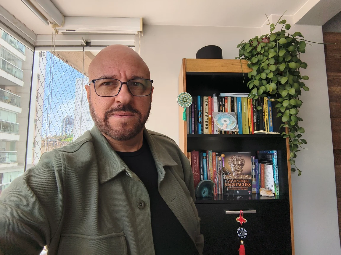 Kaique Ferreira, de óculos e camisa verde, ao lado de sua estante de livros